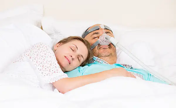 Man wearing a CPAP mask for sleep apnea treatment sleeping peacefully beside his smiling partner.